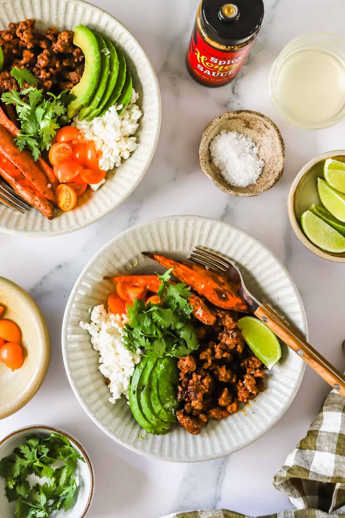Two plates with rice, sliced avocado, cooked carrots, seasoned ground meat, fresh cilantro, and lime wedges. Surrounding items include lime slices, salt, sauce, and small bowls of toppings.