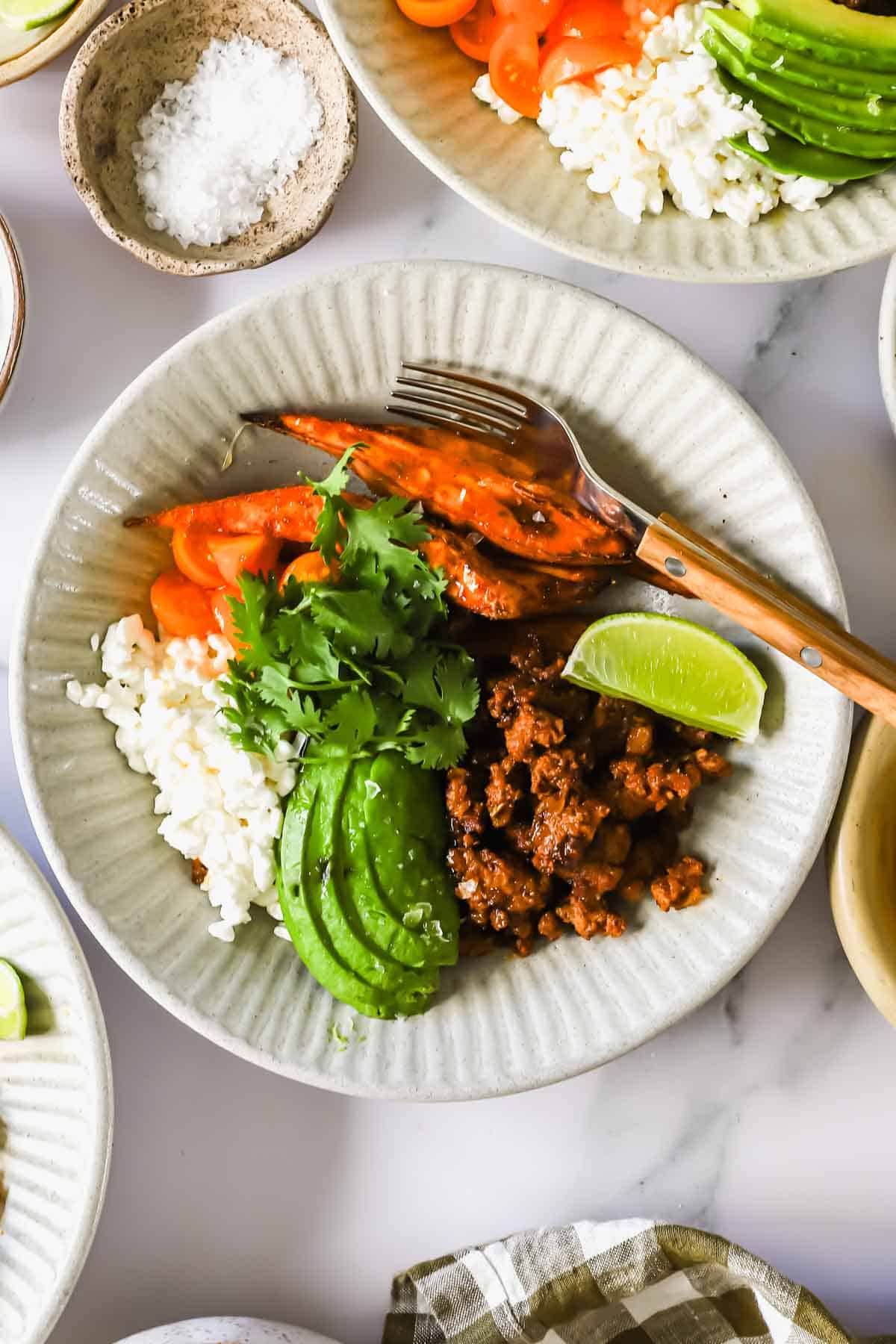 A bowl with sliced avocado, cooked ground meat, white rice, sliced carrots, cilantro, and a lime wedge, with a fork on the side.