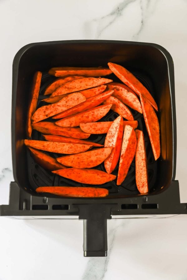 Raw sweet potato wedges coated with seasoning are arranged in a single layer inside an air fryer basket on a marble countertop.
