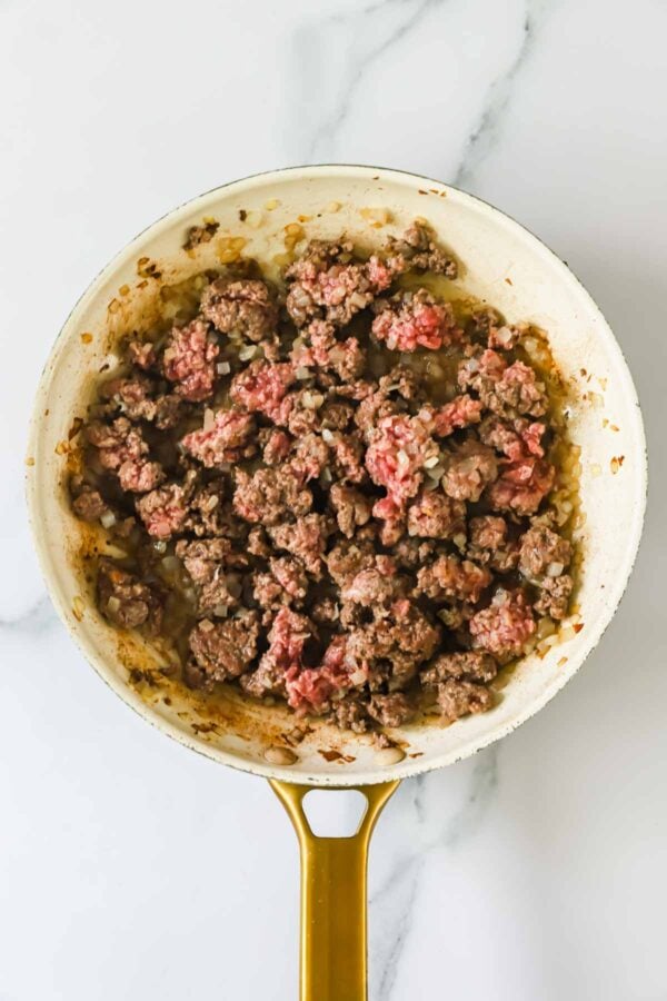Ground beef is being browned in a white skillet with a gold handle, with some pieces still partially pink and others fully cooked.