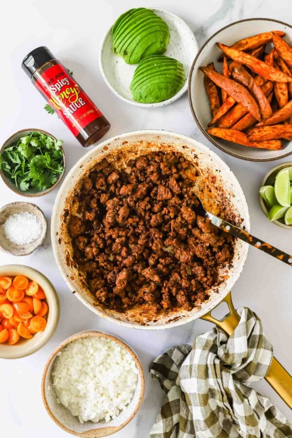 A skillet of cooked ground meat is surrounded by bowls of avocado, sweet potato fries, cilantro, lime wedges, salt, sliced carrots, rice, and a bottle of spicy seasoning.