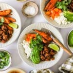 Three white plates with ground meat, rice, avocado slices, roasted sweet potatoes, tomatoes, lime wedges, and cilantro. Small bowls of salt and cilantro are nearby.
