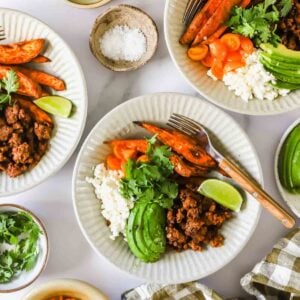 Three white plates with ground meat, rice, avocado slices, roasted sweet potatoes, tomatoes, lime wedges, and cilantro. Small bowls of salt and cilantro are nearby.