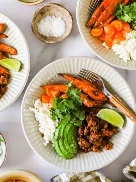 Three white plates with ground meat, rice, avocado slices, roasted sweet potatoes, tomatoes, lime wedges, and cilantro. Small bowls of salt and cilantro are nearby.
