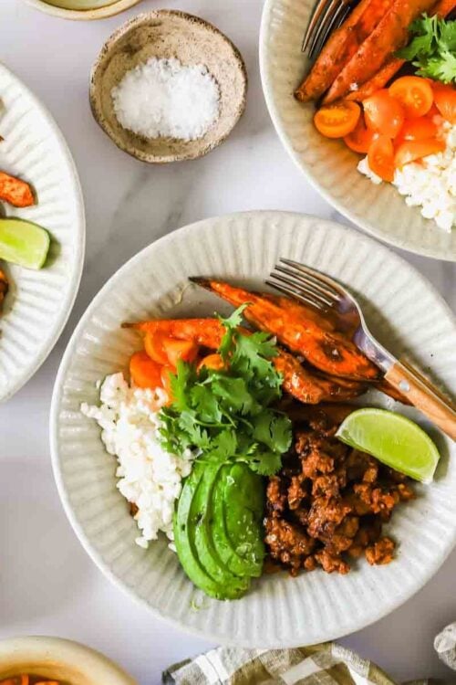 Three white plates with ground meat, rice, avocado slices, roasted sweet potatoes, tomatoes, lime wedges, and cilantro. Small bowls of salt and cilantro are nearby.
