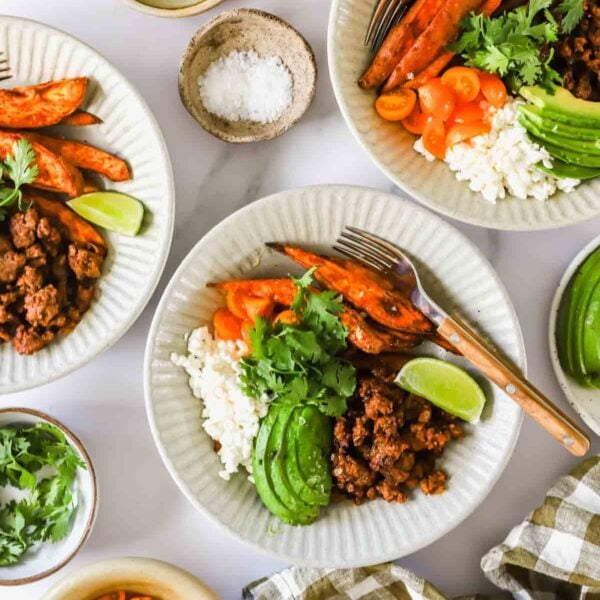 Three white plates with ground meat, rice, avocado slices, roasted sweet potatoes, tomatoes, lime wedges, and cilantro. Small bowls of salt and cilantro are nearby.