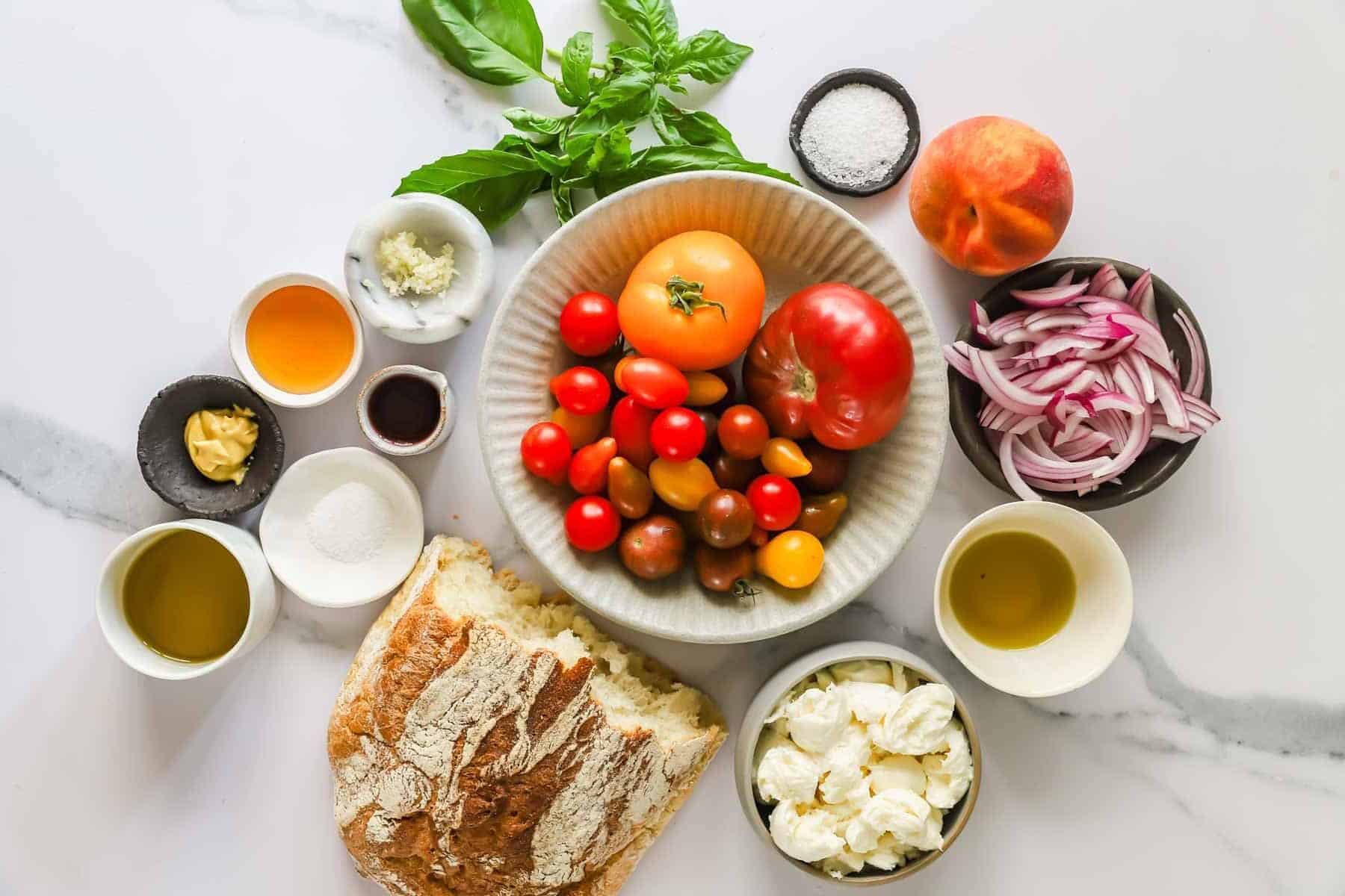 Assorted tomatoes, sliced red onion, peach, mozzarella, basil, olive oil, vinegar, mustard, garlic, salt, sugar, and rustic bread arranged on a marble surface.