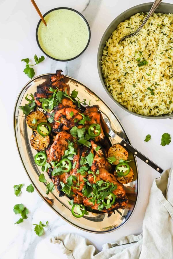Grilled chicken garnished with cilantro and jalapeños on a platter, with charred lime halves, a bowl of yellow rice, and a bowl of green sauce on a marble surface.