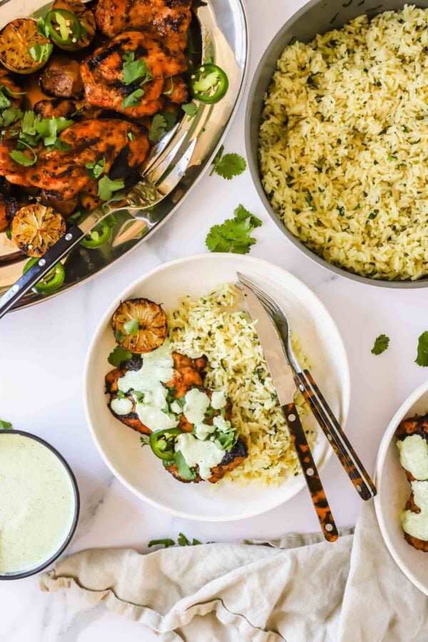 A plate with grilled chicken topped with green sauce and sliced jalapeños, rice on the side, and a bowl of rice and sauce nearby. A fork and knife are placed on the plate.