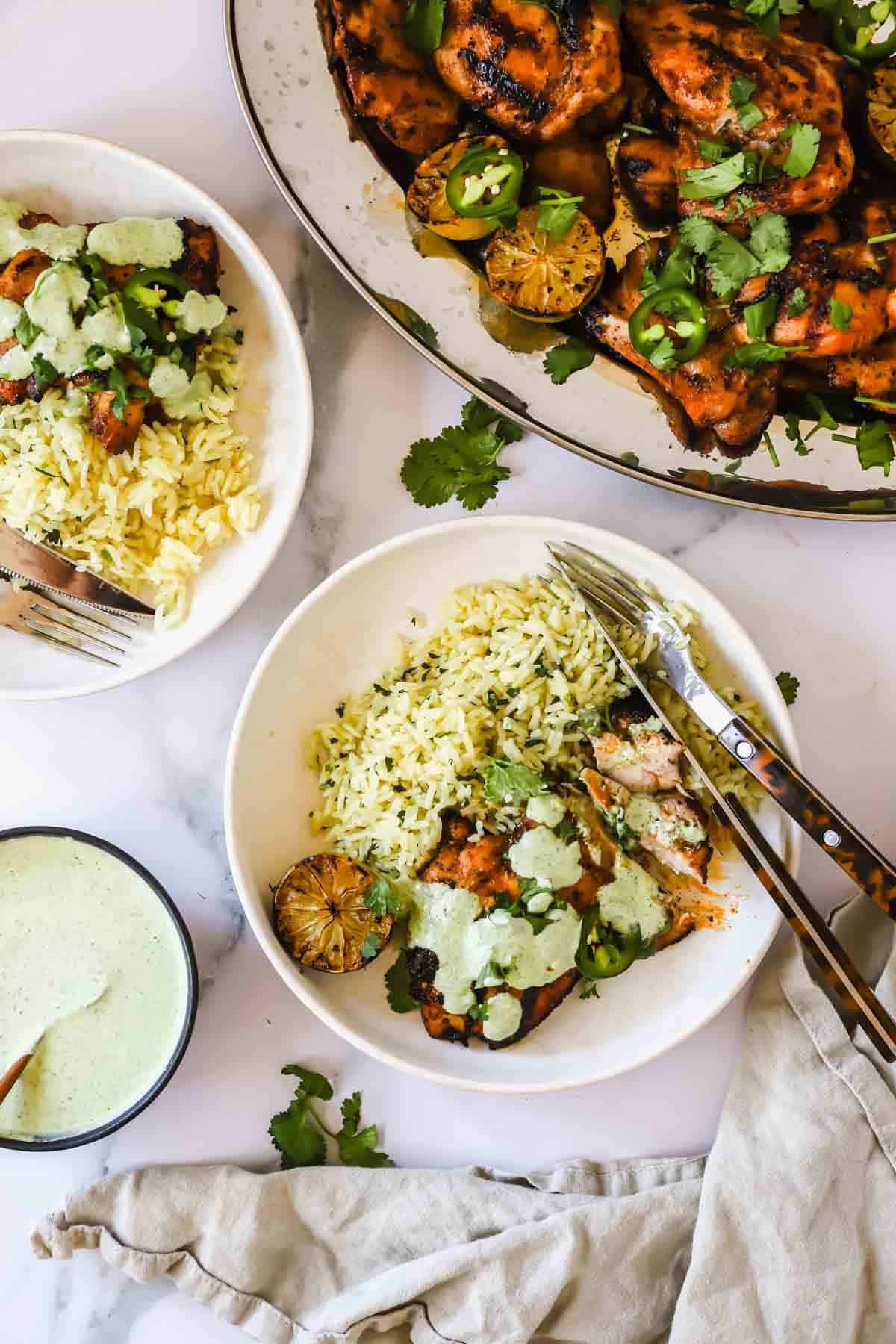 Two bowls of rice and grilled chicken topped with green sauce, garnished with cilantro and lime, next to a platter of more chicken and a small bowl of sauce on a white surface.