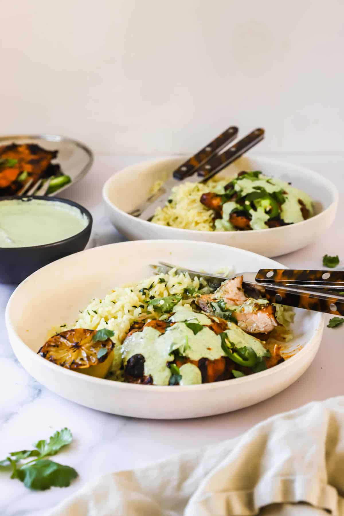 Two white bowls filled with rice, grilled chicken, roasted vegetables, and green sauce, garnished with cilantro, sit on a white surface with utensils and a small bowl of extra sauce.