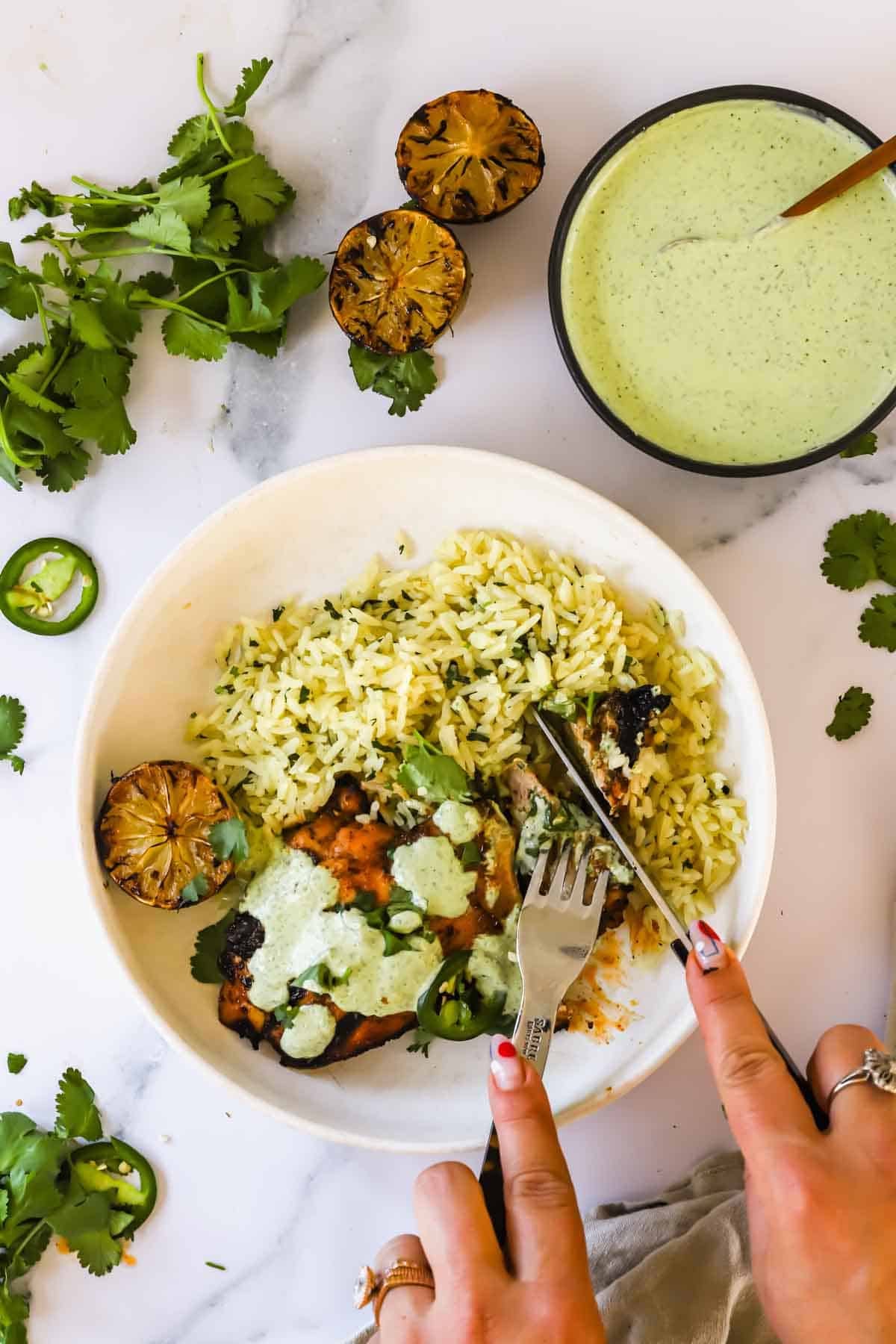 A person cuts into chicken topped with green sauce, served with herbed rice, grilled lime, and garnished with cilantro and jalapeño slices. A bowl of green sauce is nearby.