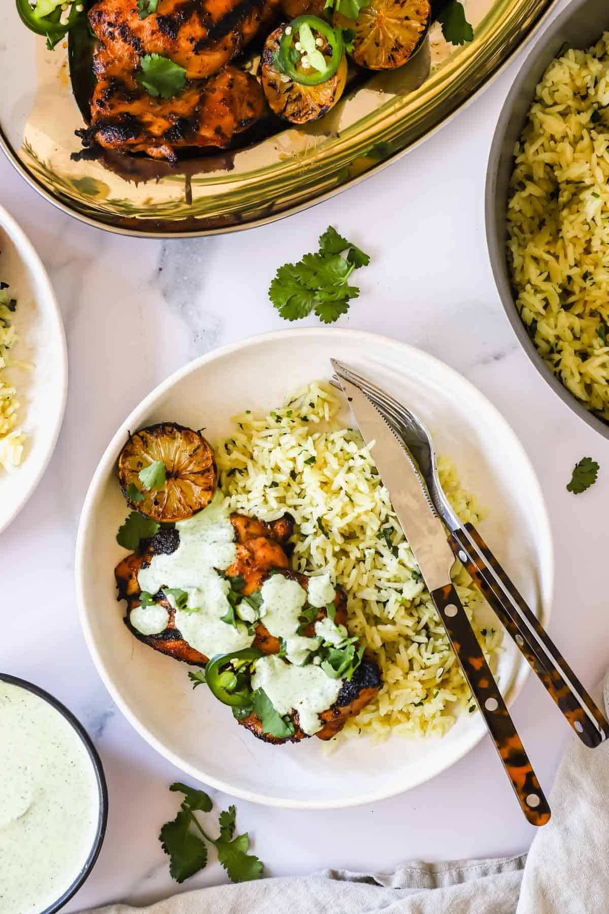 A white plate with yellow rice, grilled chicken topped with green sauce, lime slices, and cilantro, with a fork and knife on the side.