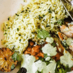 A bowl of grilled chicken topped with green sauce, yellow rice, a slice of grilled lime, and fresh herbs.