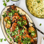 Grilled chicken garnished with cilantro and jalapeño slices on a platter with lime, served next to a bowl of seasoned rice and a small bowl of green sauce.