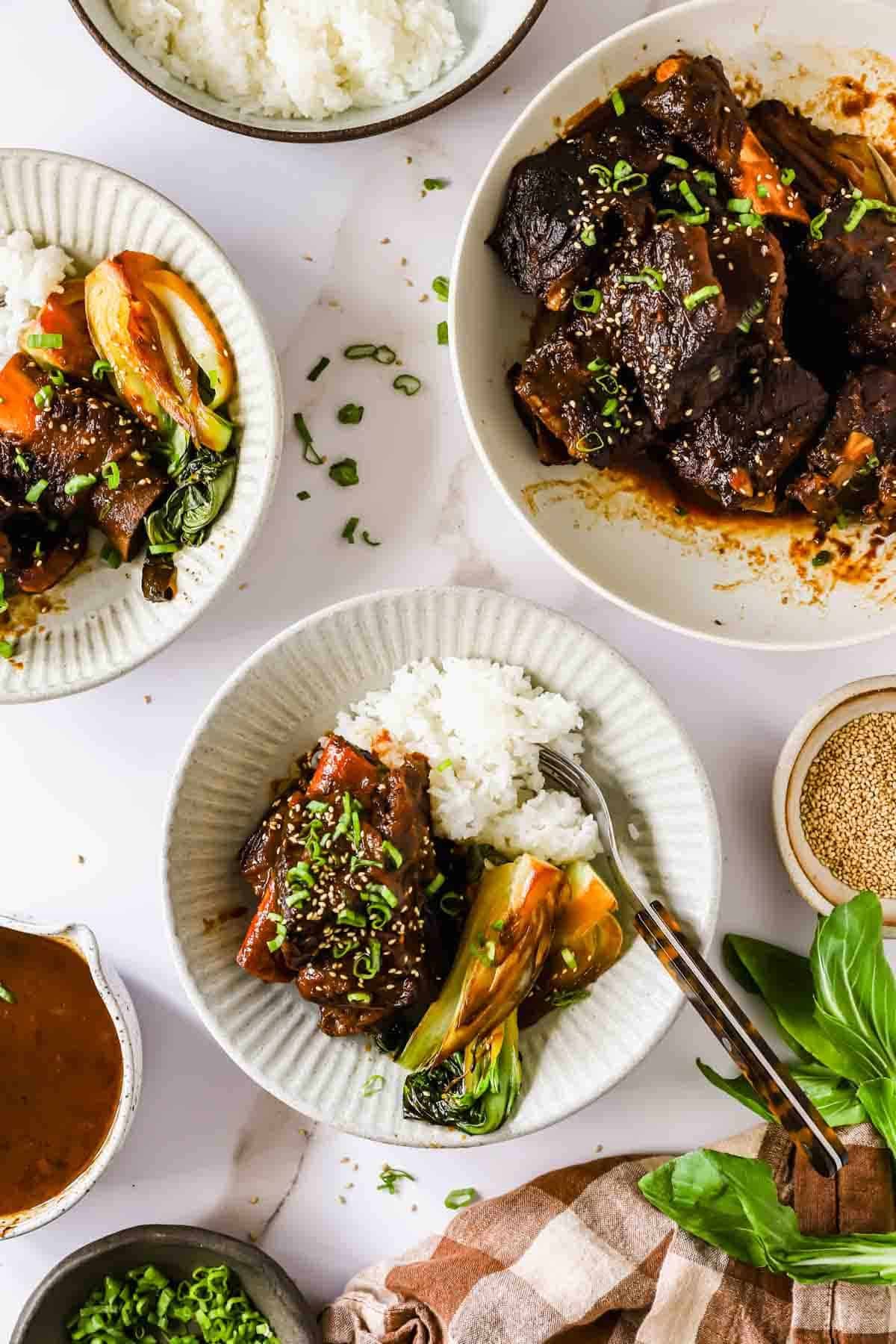 Bowls of white rice topped with braised short ribs, bok choy, carrots, and green onions, with extra meat and sauce served on the side.
