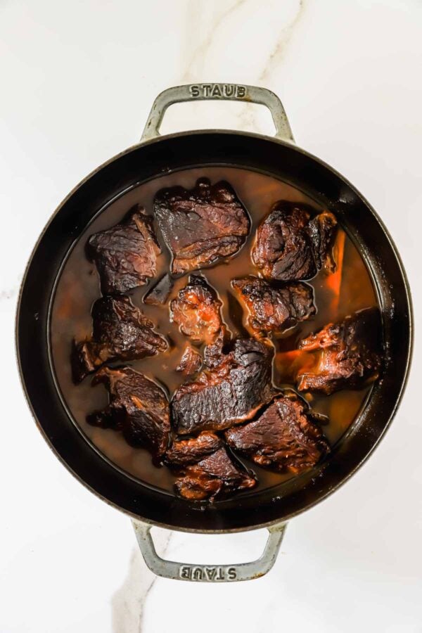 Braised beef short ribs in a round cast iron pot with dark broth, viewed from above on a white surface.