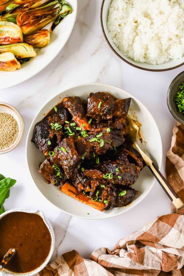 Braised short ribs garnished with green onions served in a white bowl, with sides of rice, sautéed bok choy, sesame seeds, and sauce on a marble surface.