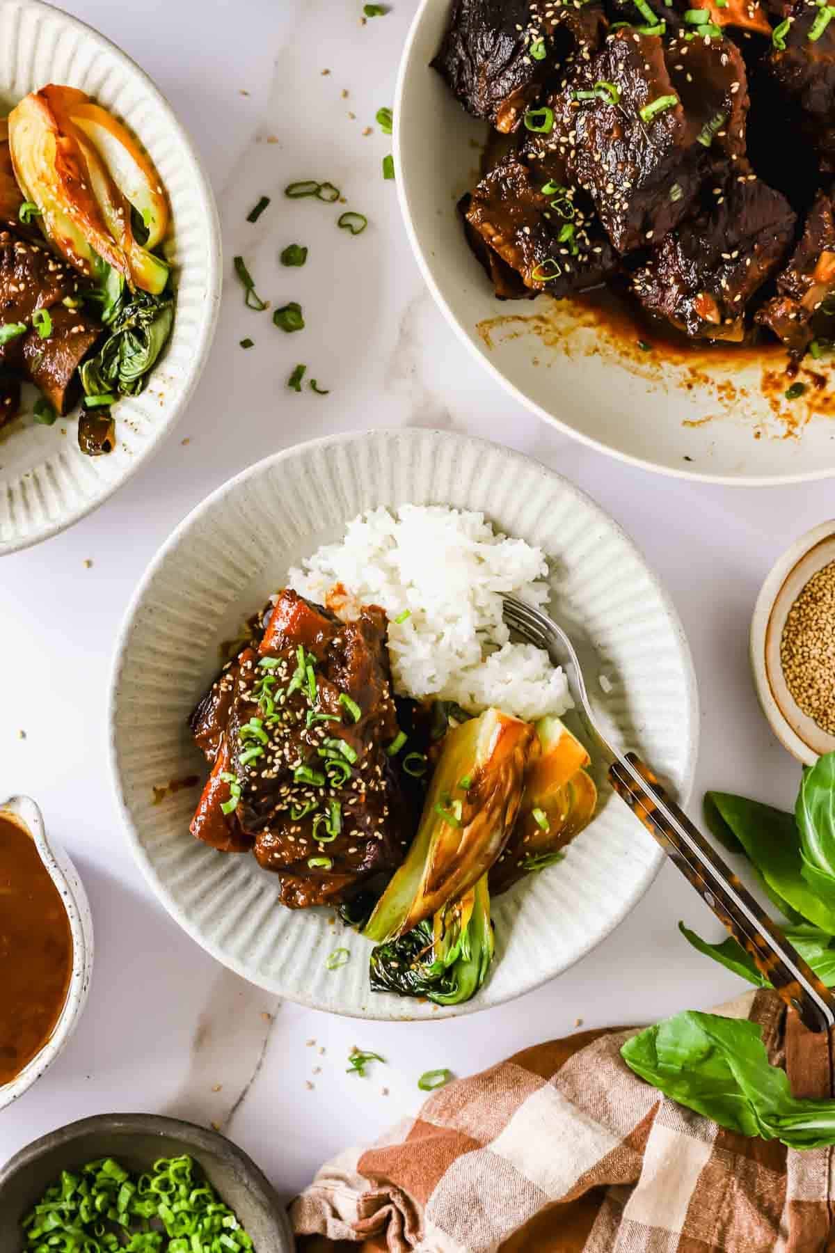 A plate with braised short ribs, white rice, and bok choy, garnished with chopped green onions, sits on a table with similar dishes and fresh herbs nearby.