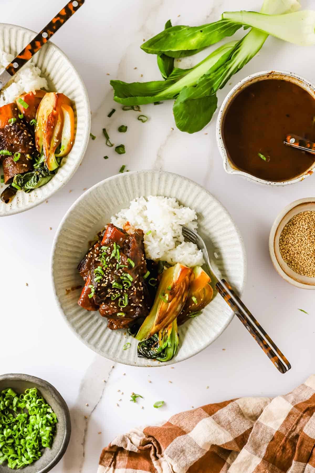 A plate with white rice, glazed meat, and bok choy, garnished with green onions. Surrounding items include extra sauce, sesame seeds, sliced green onions, and fresh bok choy on a marble surface.