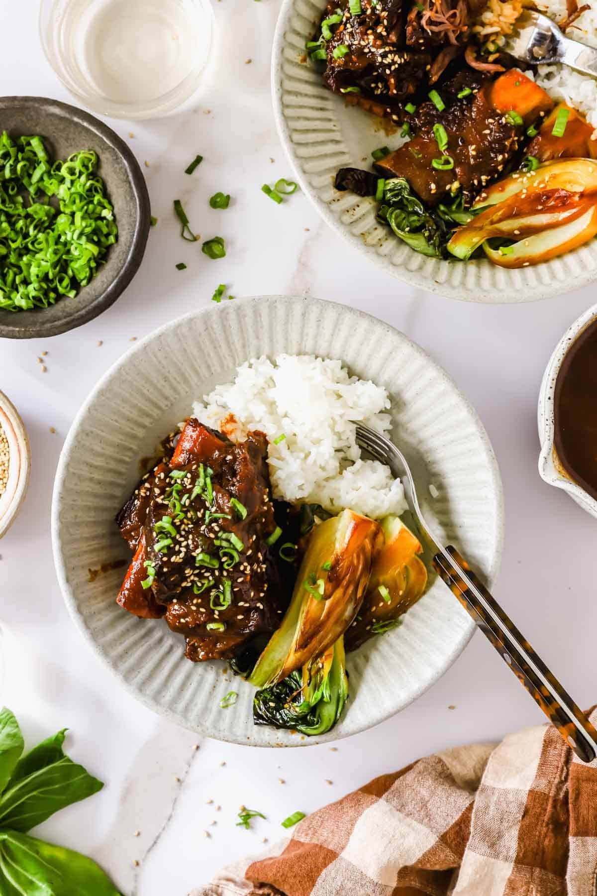 A plate with white rice, glazed short ribs, bok choy, and sliced green onions, next to a fork and a bowl of chopped green onions.