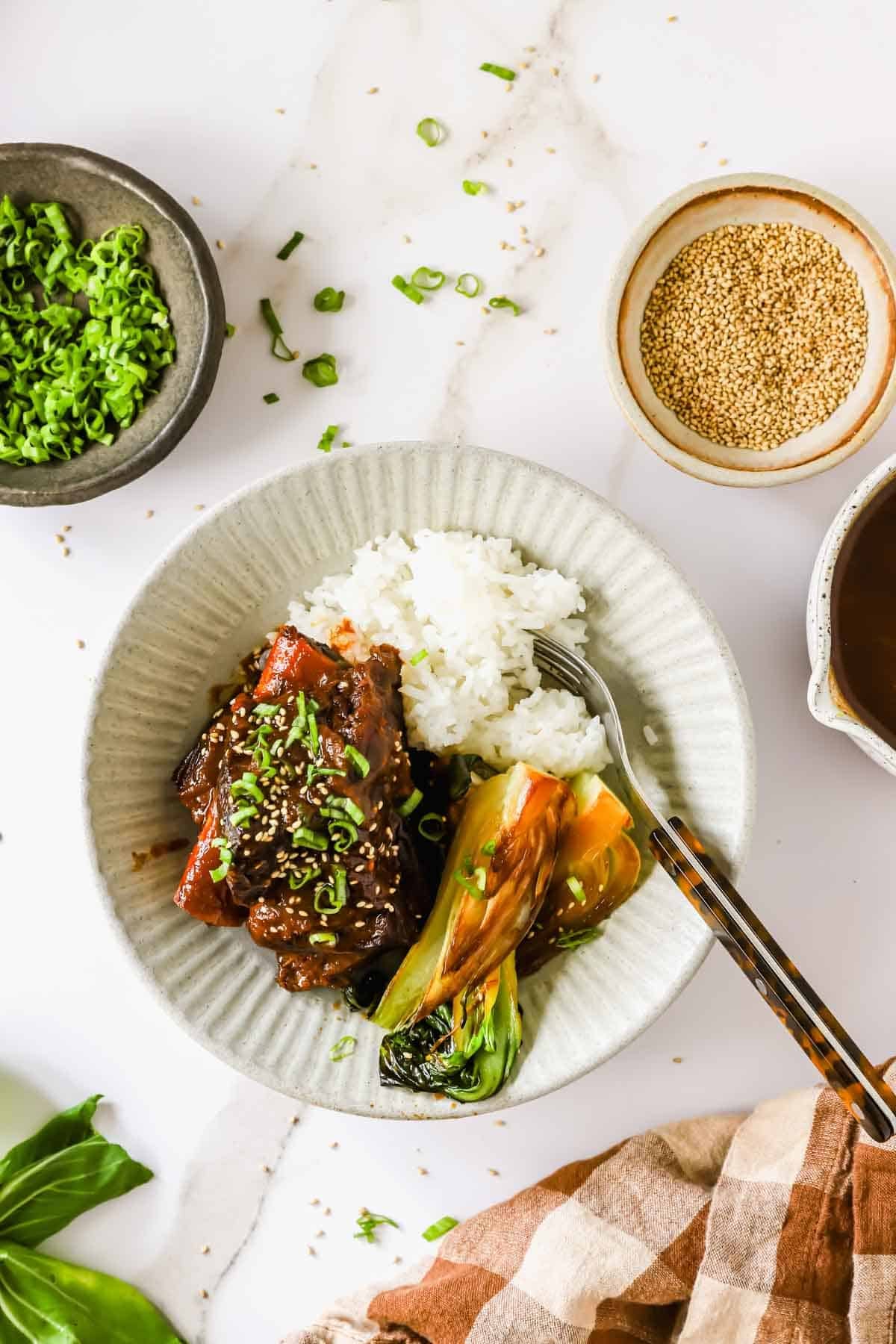A plate with white rice, glazed meat, and bok choy, garnished with sliced green onions, beside bowls of green onions, sesame seeds, and sauce on a white surface.