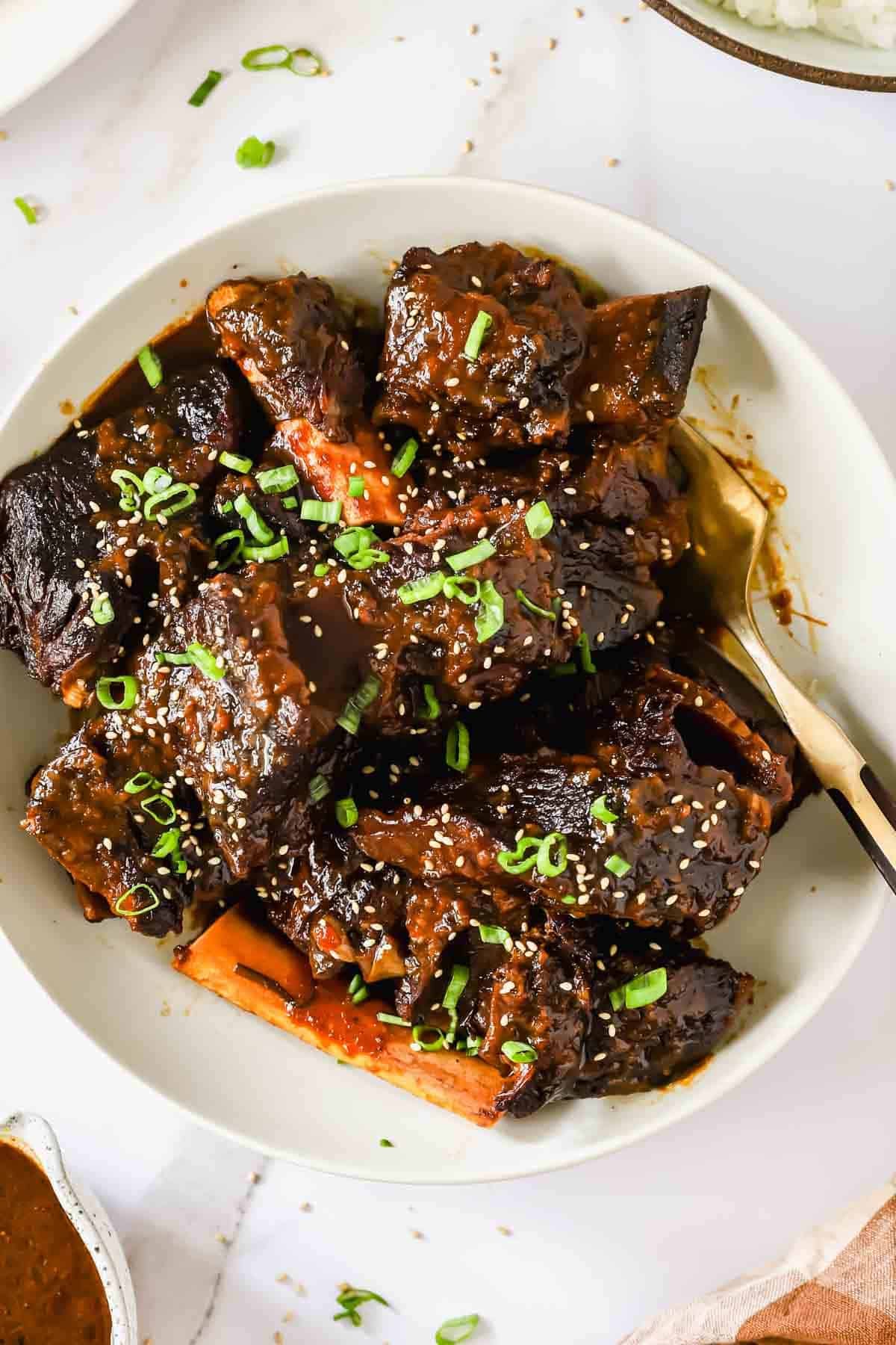 Bowl of braised beef short ribs in dark sauce, topped with chopped green onions and sesame seeds, with a fork on the side.