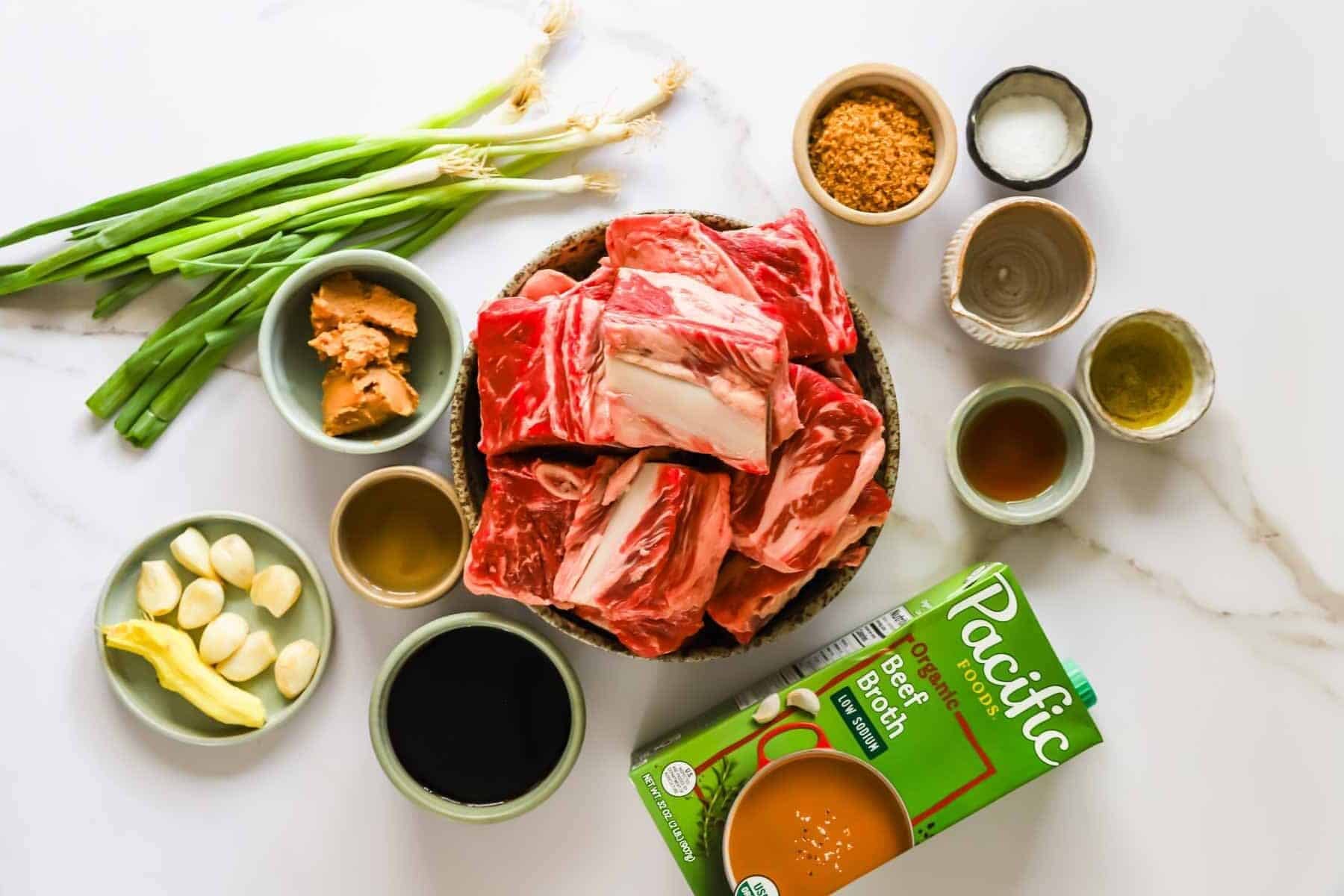 Raw short ribs in a bowl surrounded by green onions, garlic, ginger, brown sugar, soy sauce, miso paste, sesame oil, salt, and a carton of beef broth on a marble surface.