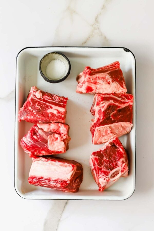 Six browned beef short ribs are arranged in a round black cast iron pot on a white marble surface.