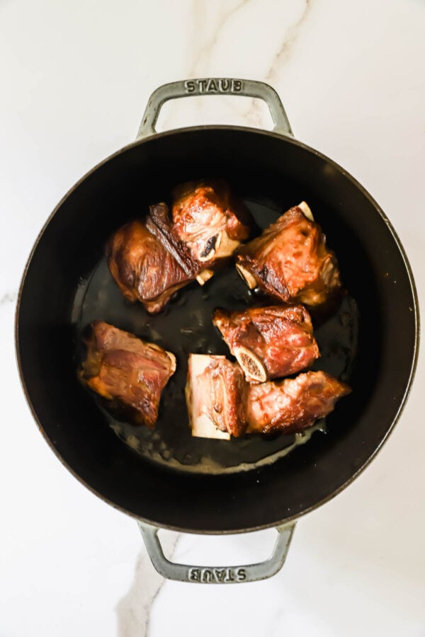 Six browned beef short ribs are arranged in a round black cast iron pot on a white marble surface.