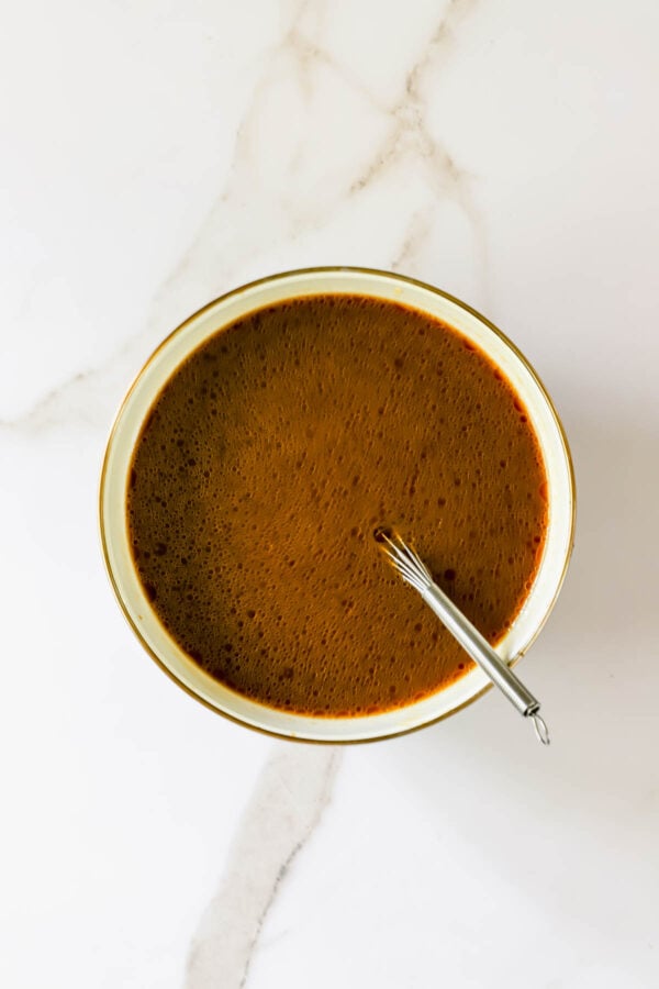 A bowl filled with a brown liquid, possibly soup or sauce, with a small metal whisk resting inside on a white marble surface.