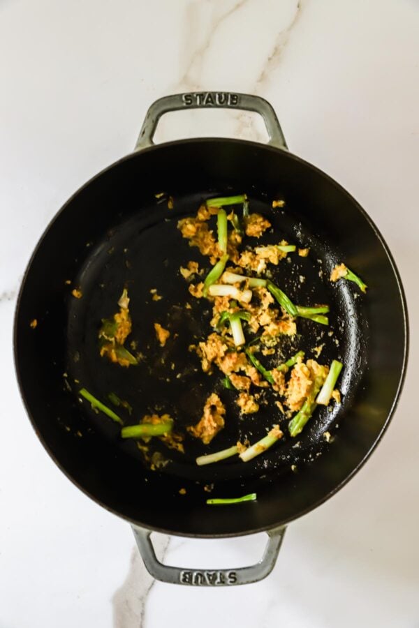 Black pot on a white surface containing partially cooked ground meat and sliced green onions, with some residue visible on the bottom of the pot.