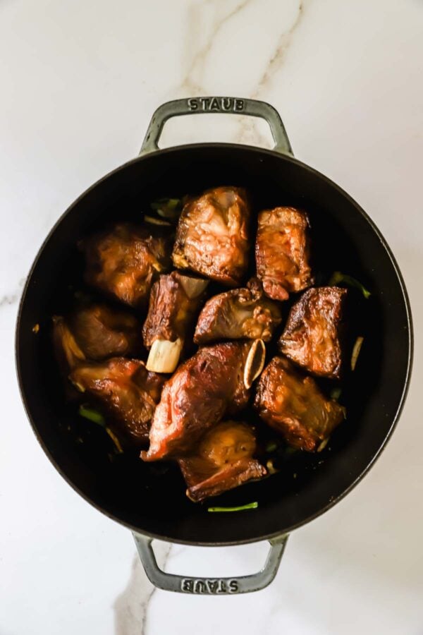 Braised beef short ribs are arranged in a black Dutch oven on a white marble surface.