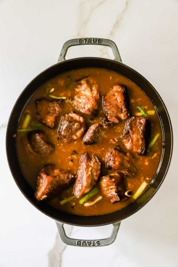 A black pot filled with braised beef short ribs in brown sauce, garnished with green onions, on a white marble surface.