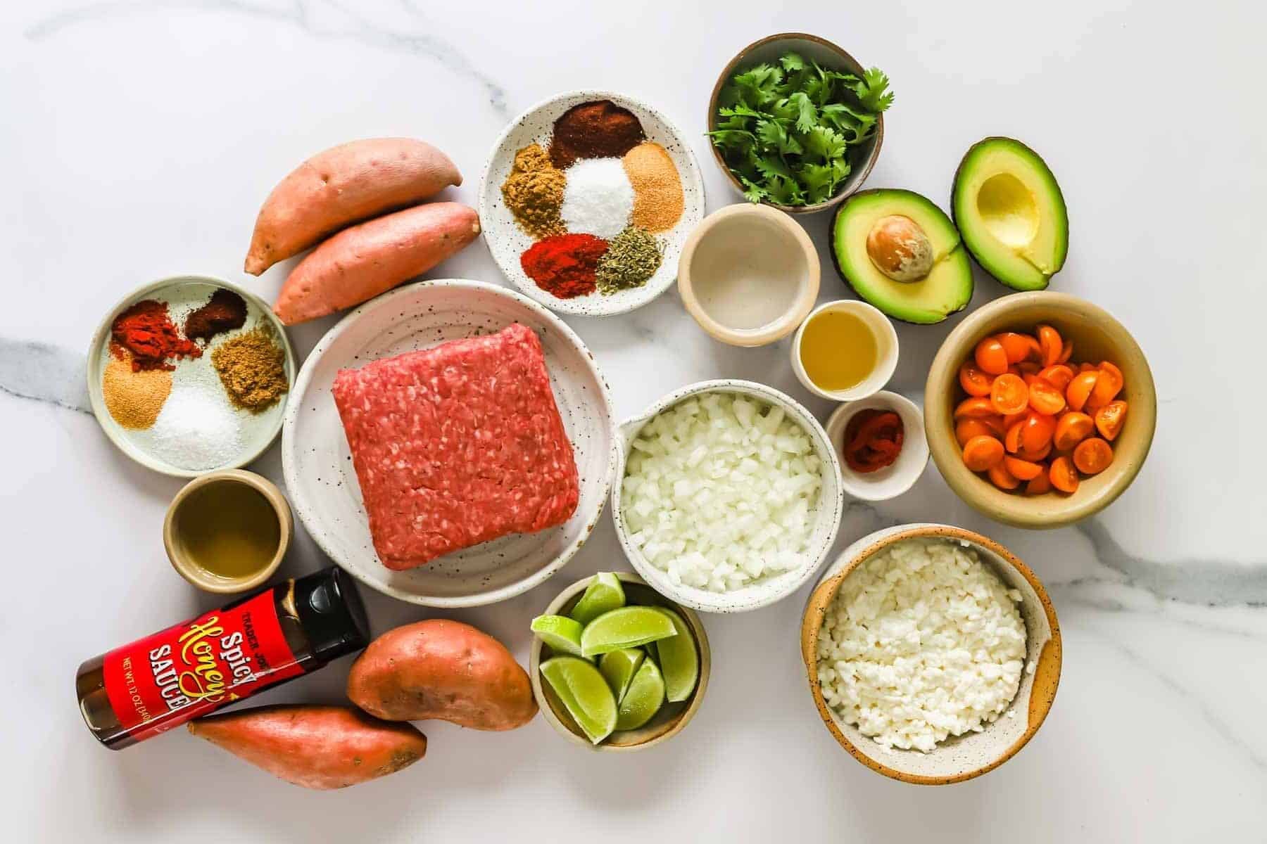 Assorted ingredients on a white surface, including ground meat, sweet potatoes, rice, onions, lime wedges, avocado, tomatoes, spices, cilantro, oil, and a bottle of spicy honey sauce.