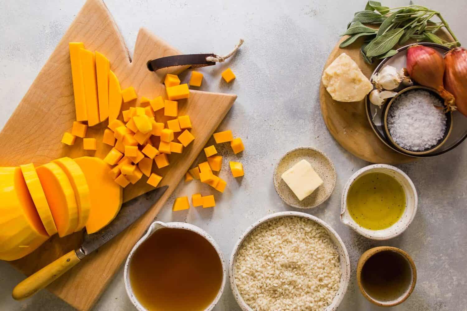 Chopping butternut squash on a cutting board. Rice, broth, butter, oil, and cheese on the side.