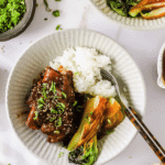 A bowl with braised beef short ribs, white rice, bok choy, and chopped green onions, garnished with sesame seeds, placed on a white surface.