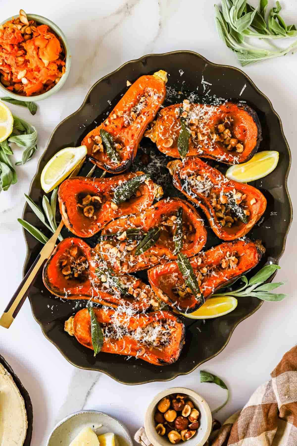 Roasted butternut squash halves topped with hazelnuts, sage, and grated cheese, served on a platter with lemon wedges.