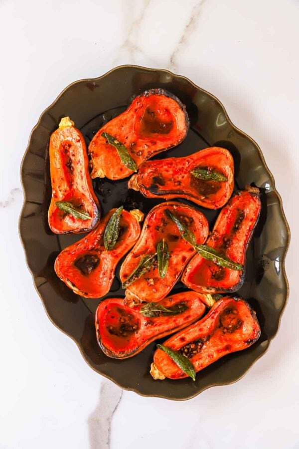 Roasted butternut squash halves topped with fried sage leaves on a dark platter.