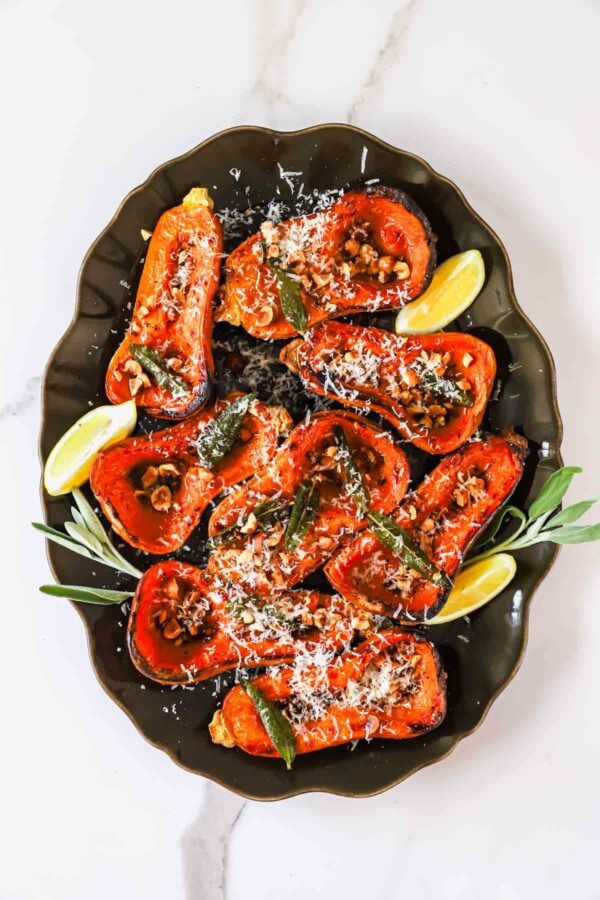 Roasted butternut squash halves topped with nuts, sage, and grated cheese on a platter with lemon wedges.