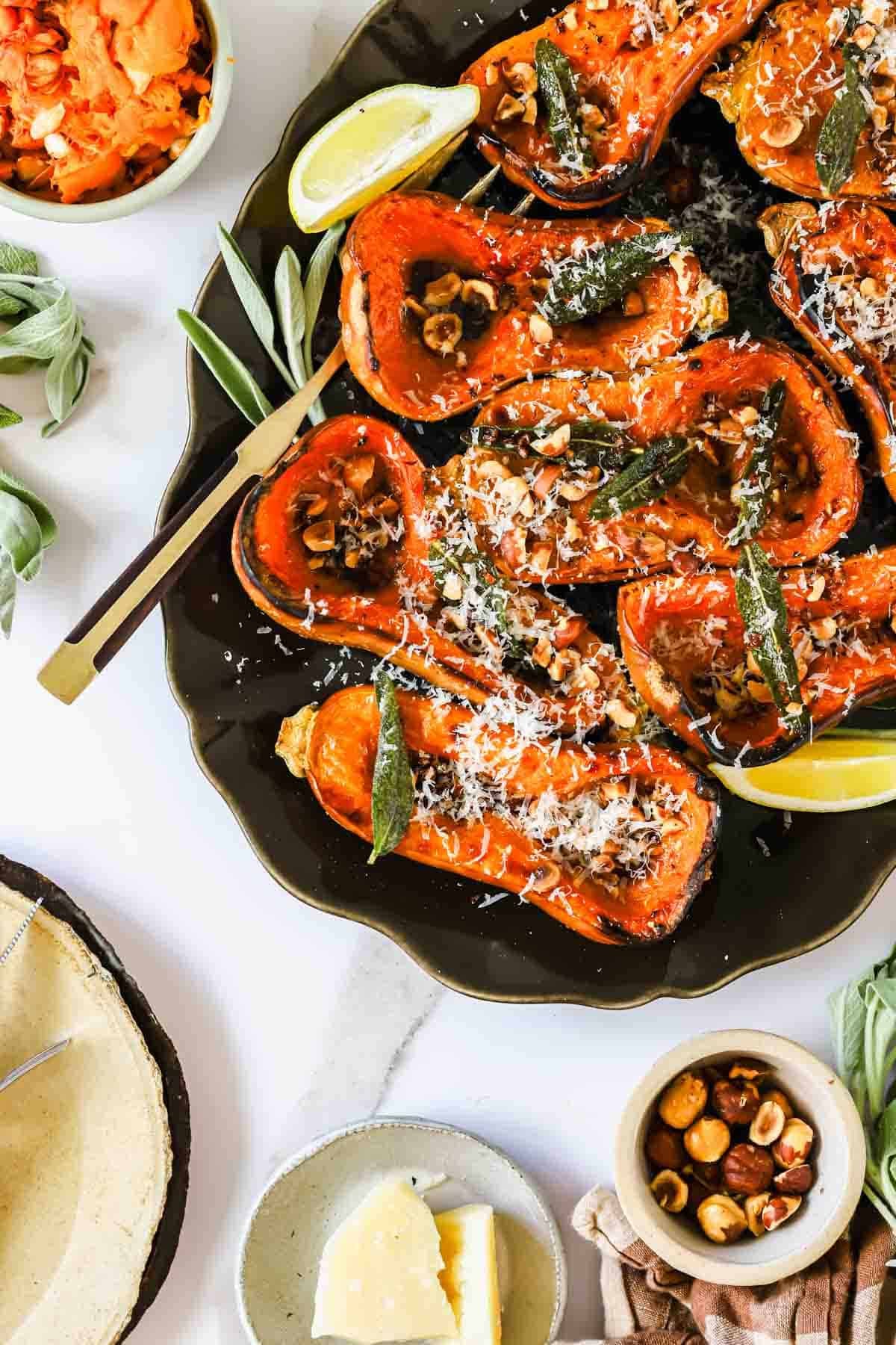 Roasted butternut squash halves topped with nuts, sage leaves, and grated cheese on a platter with lemon wedges.