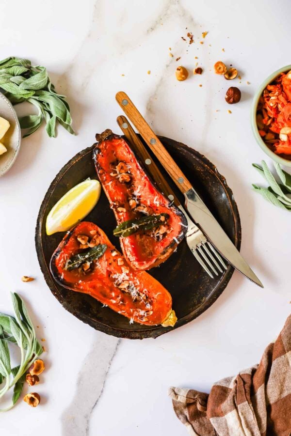 Two roasted butternut squash halves topped with nuts, herbs, and cheese, served on a dark plate with a lemon wedge, fork, and knife.