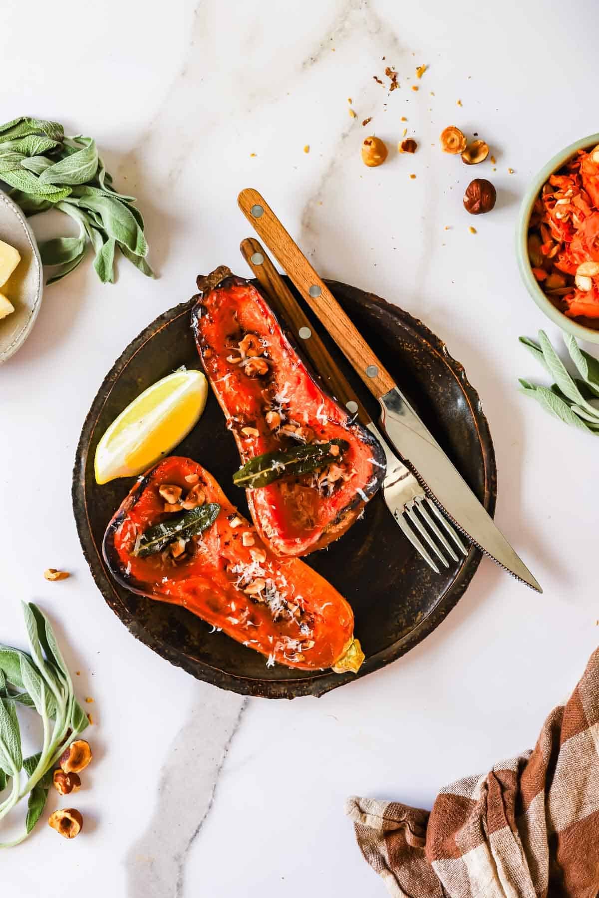 Two roasted butternut squash halves topped with nuts, herbs, and cheese, served on a dark plate with a lemon wedge, fork, and knife.