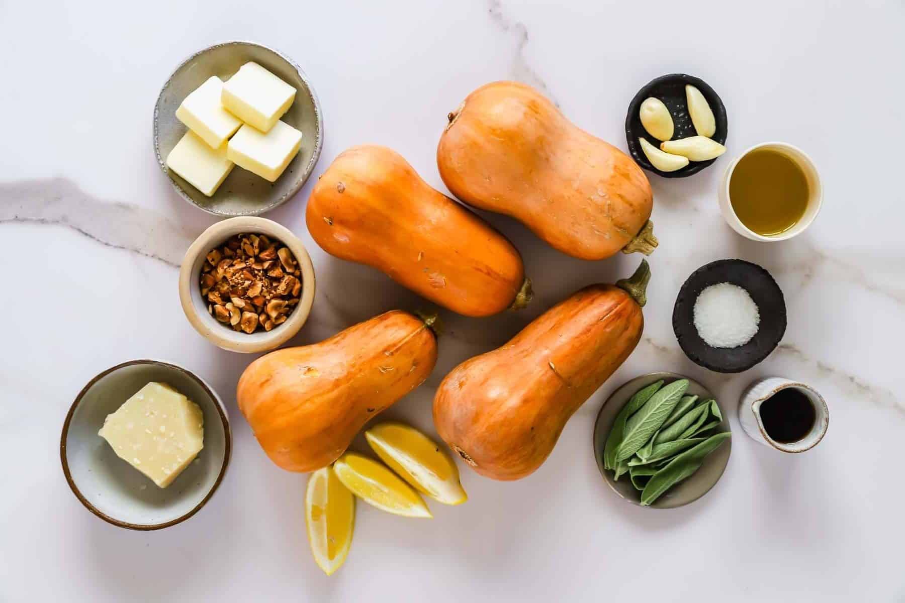 Four butternut squash arranged with bowls containing butter, garlic, sage, nuts, oil, salt, balsamic vinegar, parmesan, and lemon wedges.