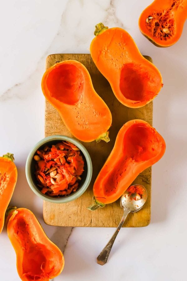 Butternut squash halved and scooped out on a wooden cutting board, with a small bowl of seeds and pulp and a silver spoon.