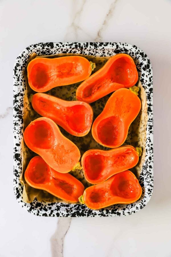 Eight halved butternut squash arranged in a rectangular speckled baking pan.