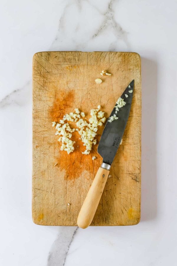 A chef’s knife resting on a wooden cutting board next to a pile of minced garlic.