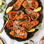 Roasted butternut squash halves topped with nuts, herbs, and grated cheese on a platter with lemon wedges.