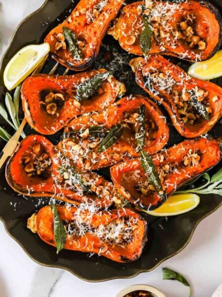 Roasted butternut squash halves topped with nuts, herbs, and grated cheese on a platter with lemon wedges.