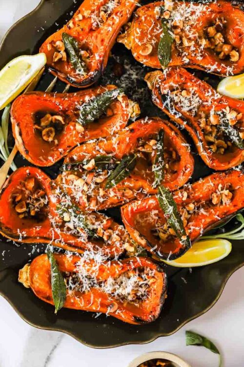 Roasted butternut squash halves topped with nuts, herbs, and grated cheese on a platter with lemon wedges.
